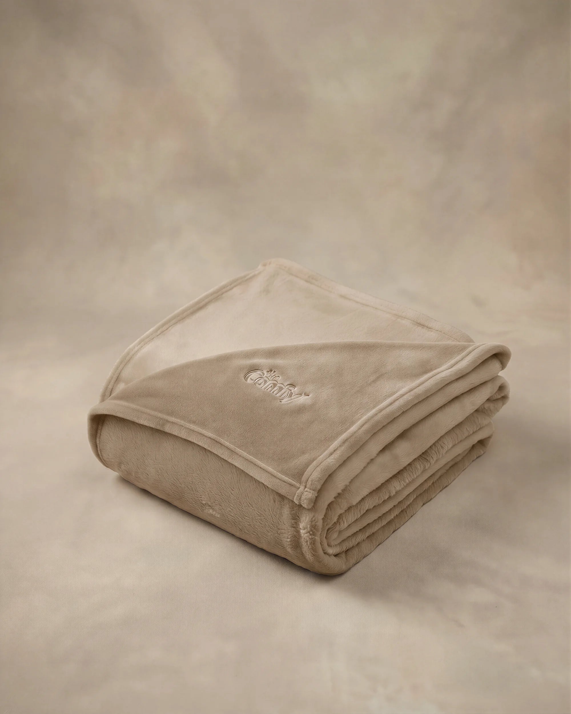 Folded tan oversized blanket with embroidered logo displayed on a matching neutral backdrop.