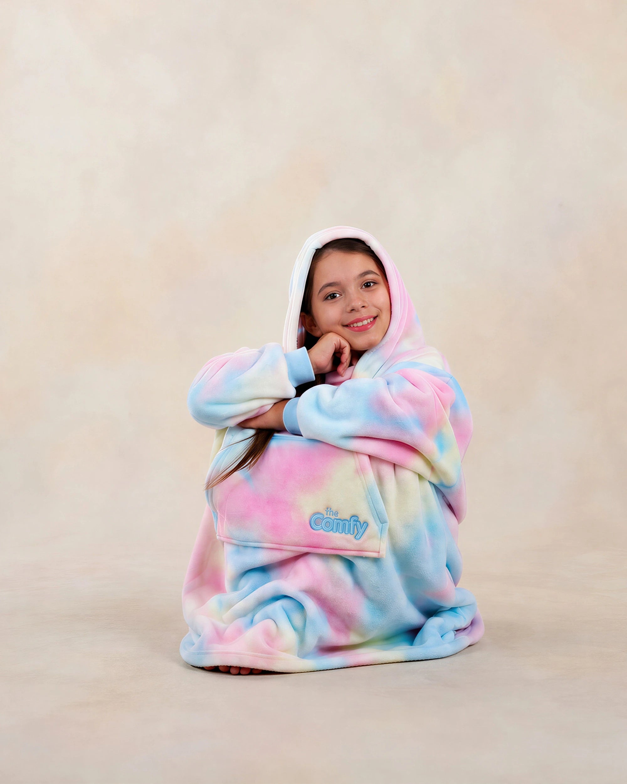Girl sitting comfortably in a pastel tie-dye wearable blanket with her chin resting on her hand against a neutral studio background.