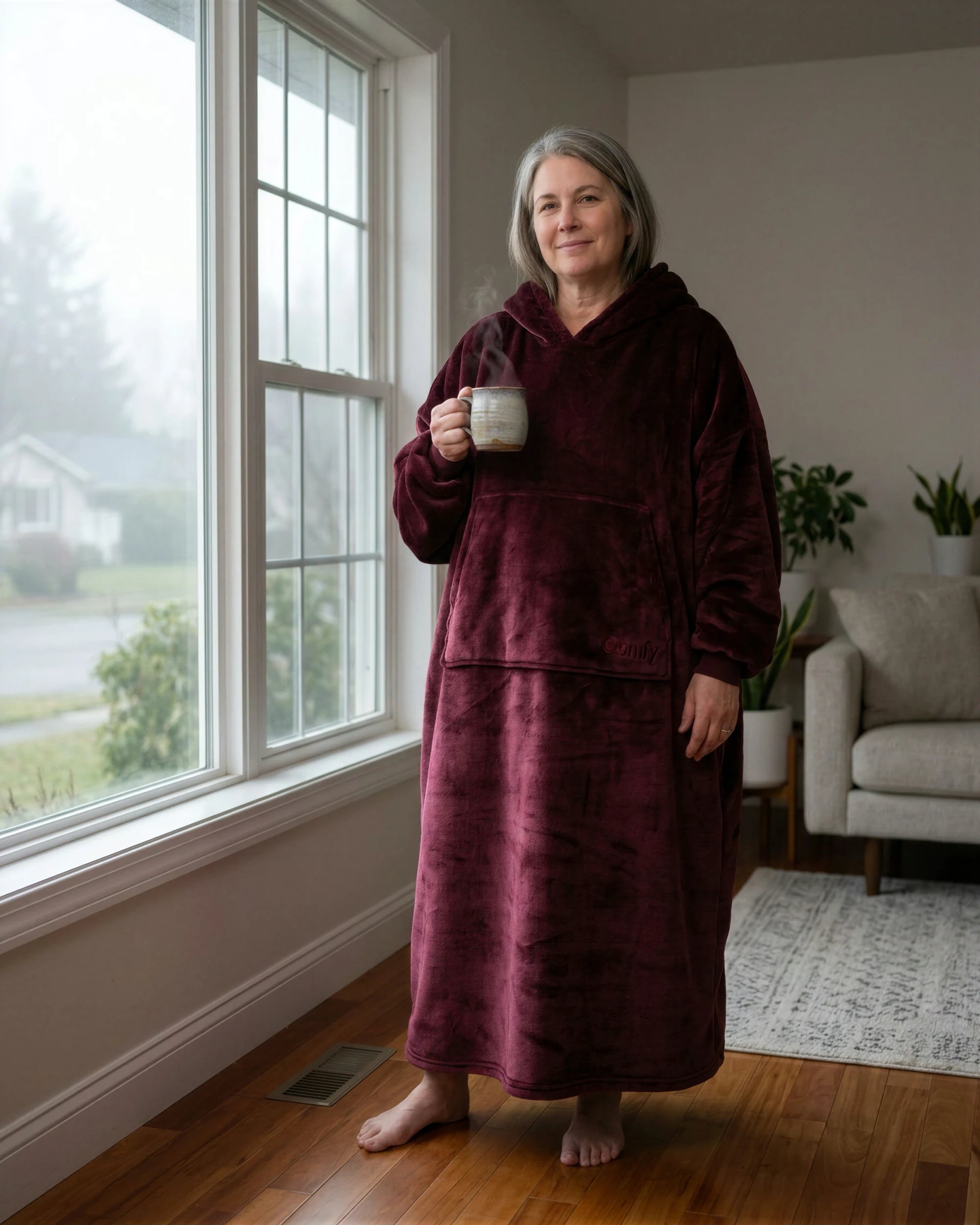 Woman standing by a window holding a mug while wearing The Dream Long in Burgundy in a cozy home setting.