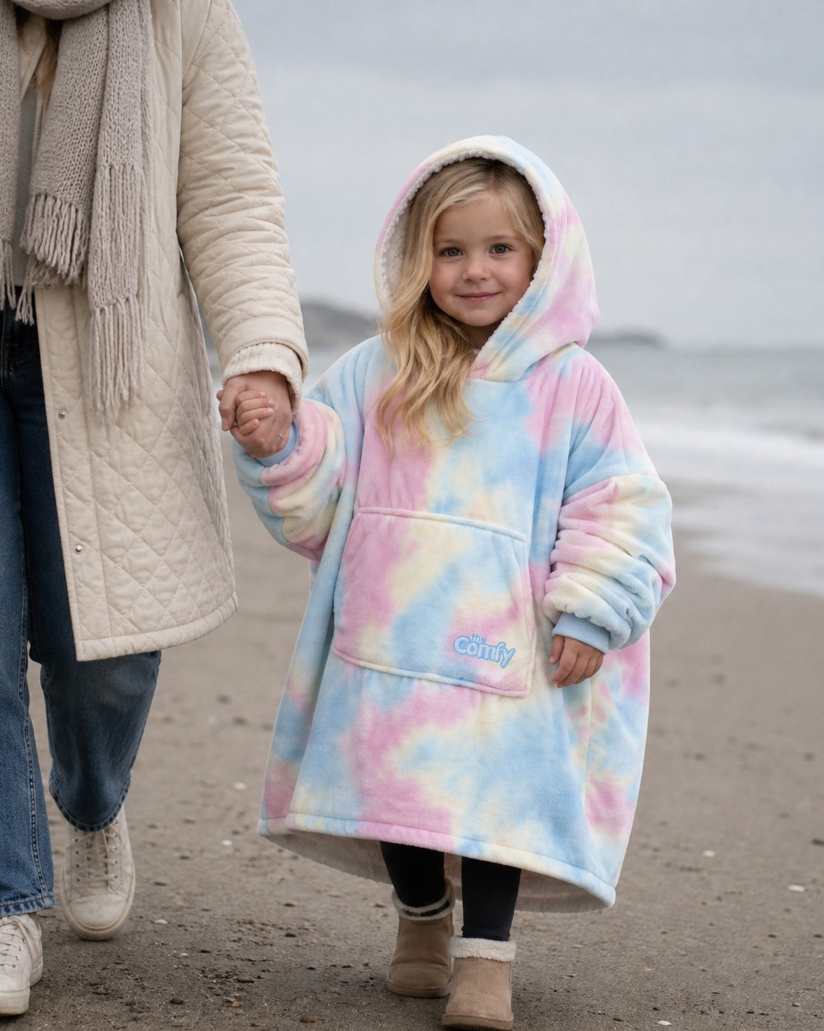 A young girl walking on a beach while holding an adult's hand, wearing The Original Jr. wearable blanket in Cotton Candy. The hoodie features soft pink, blue, and yellow tie-dye patterns with a front kangaroo pocket and a small embroidered 'The Comfy' logo.