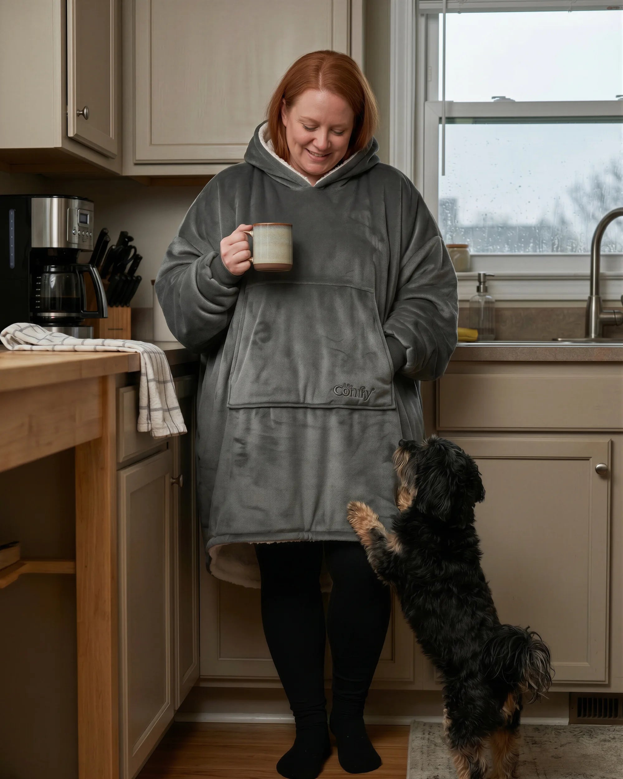 Woman wearing The Original wearable blanket in charcoal holding a mug in a kitchen with a dog nearby