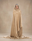 Woman wrapped head-to-toe in a tan oversized blanket, standing against a neutral beige studio backdrop.