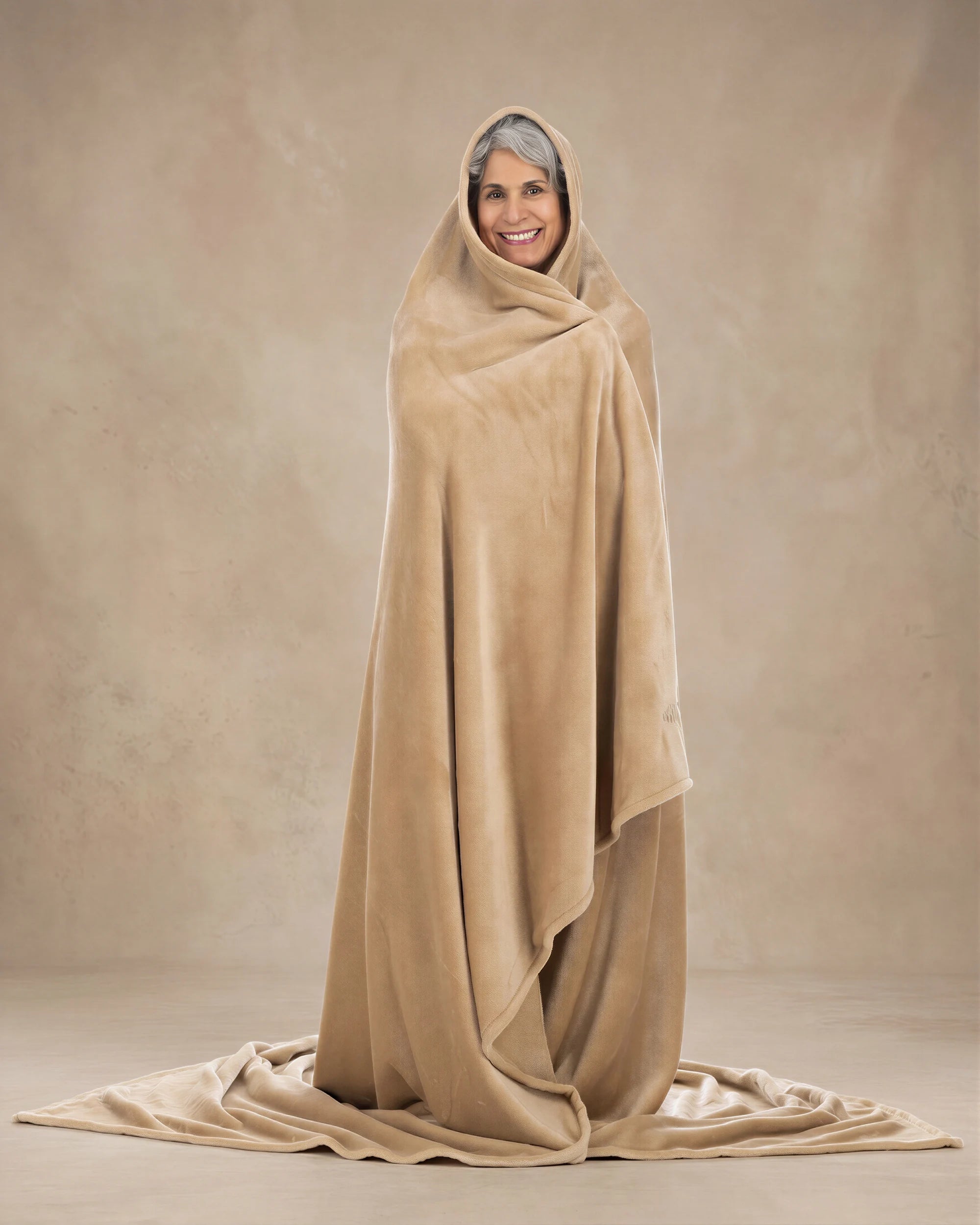 Woman wrapped head-to-toe in a tan oversized blanket, standing against a neutral beige studio backdrop.