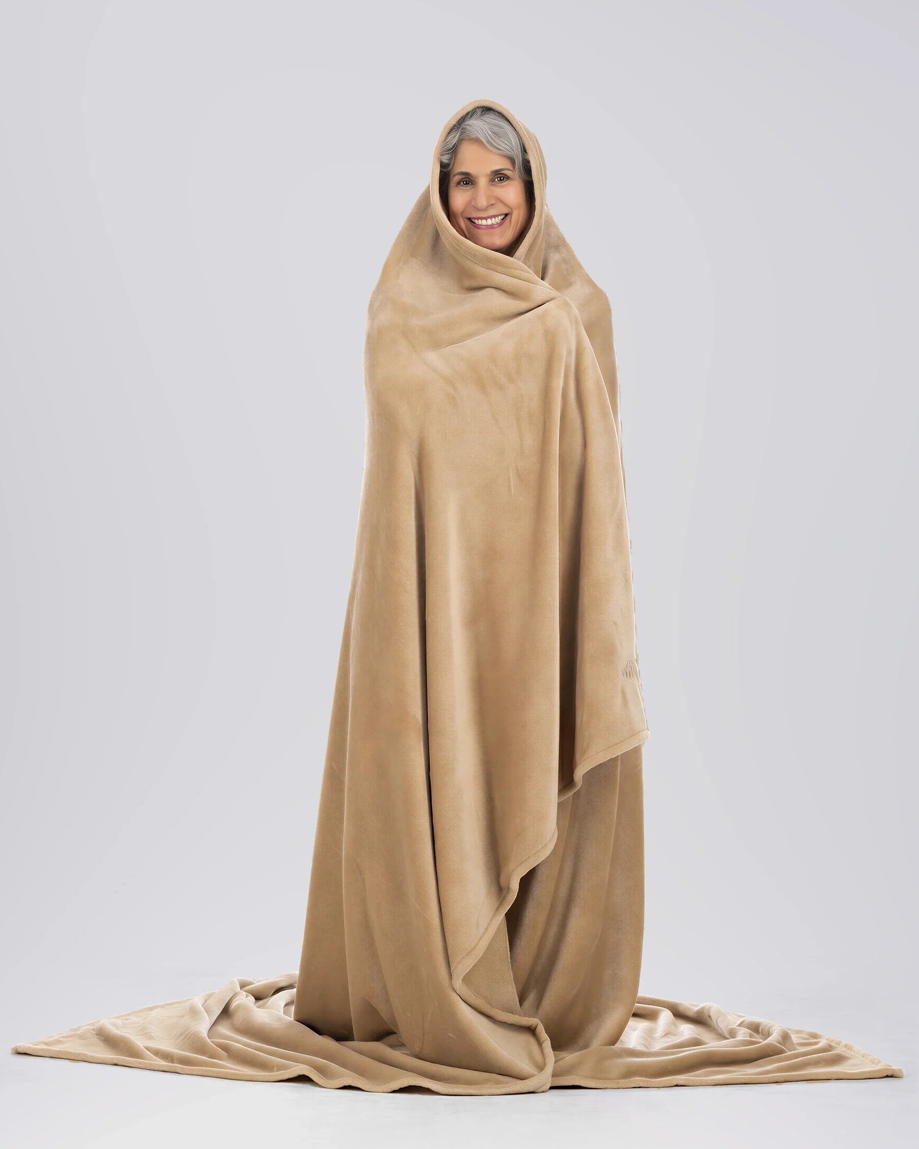 Woman wrapped head-to-toe in a tan oversized blanket, standing against a neutral beige studio backdrop.