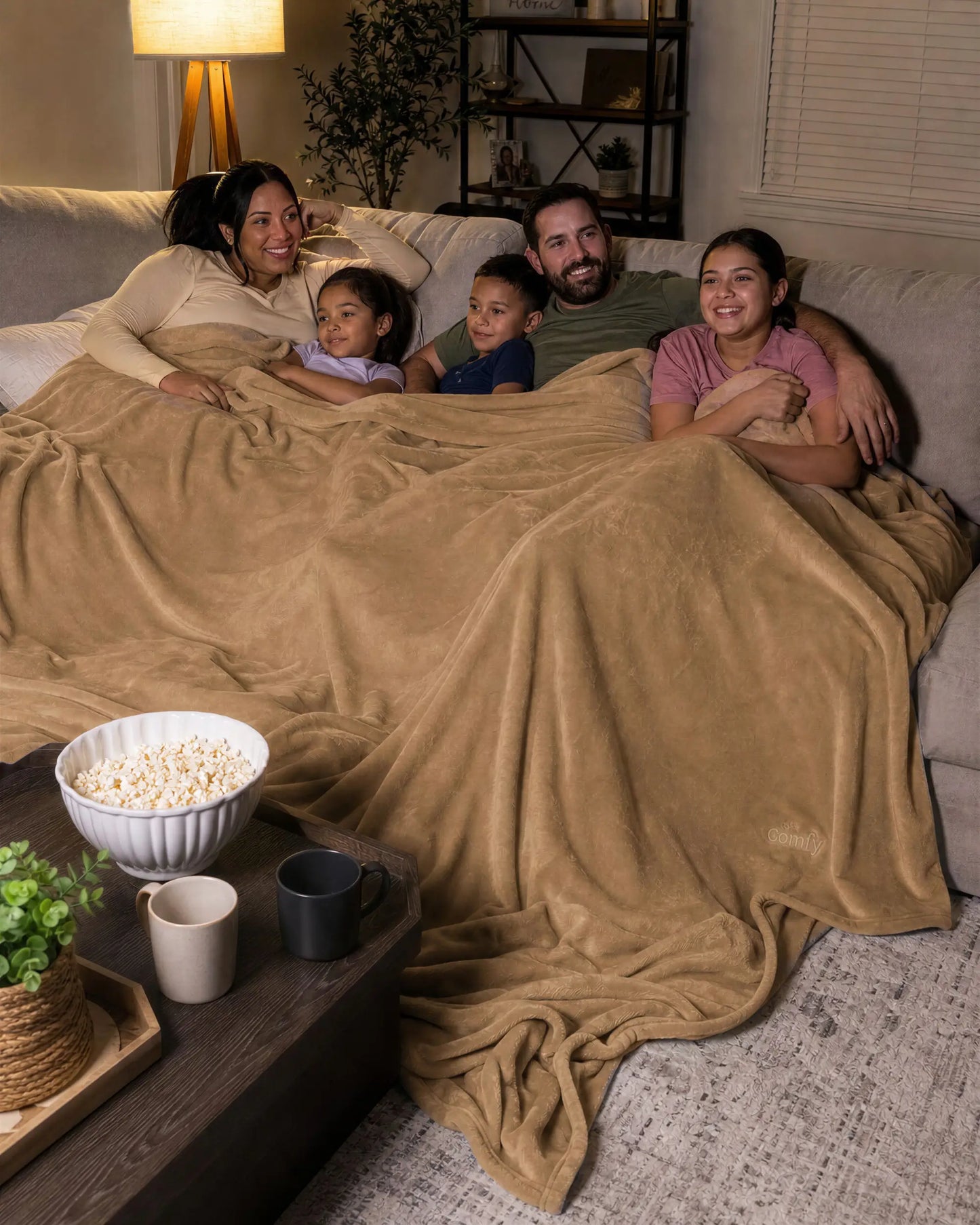 Family of five relaxing together on a sectional sofa under a tan oversized blanket during a cozy movie night.