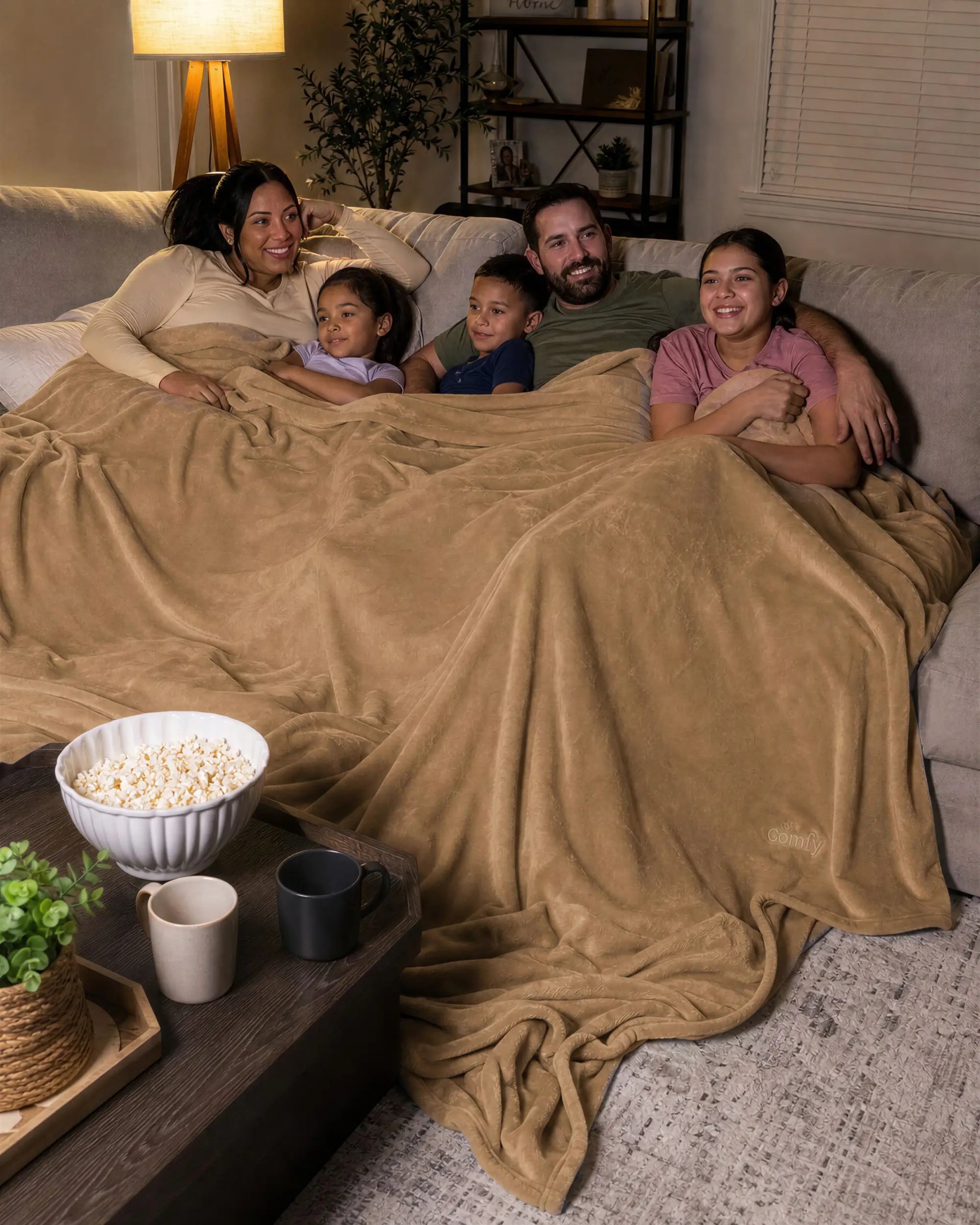 Family of five relaxing together on a sectional sofa under a tan oversized blanket during a cozy movie night.