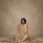 Woman sitting on the floor wrapped in a tan oversized blanket, smiling against a neutral studio background.