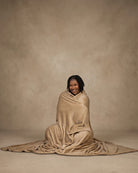 Woman sitting on the floor wrapped in a tan oversized blanket, smiling against a neutral studio background.