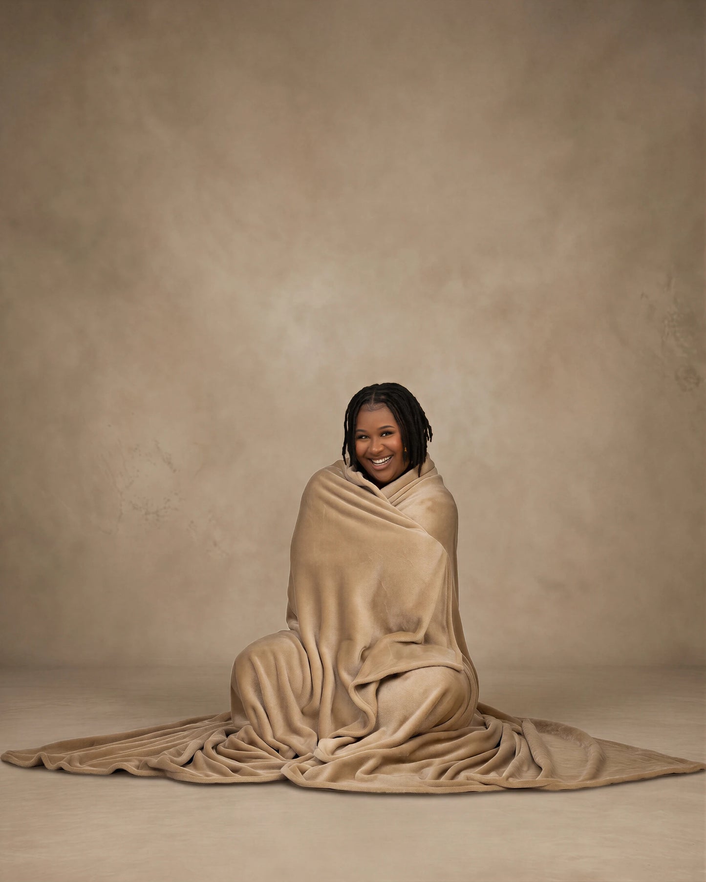 Woman sitting on the floor wrapped in a tan oversized blanket, smiling against a neutral studio background.