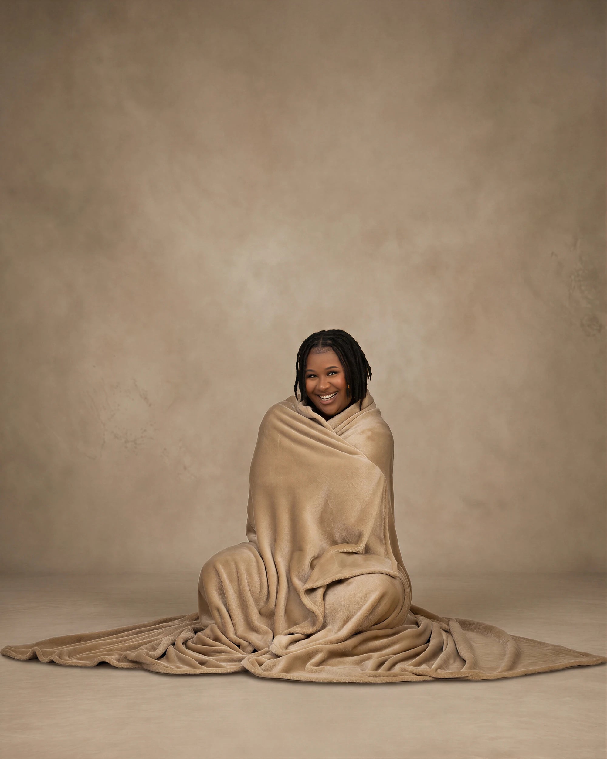 Woman sitting on the floor wrapped in a tan oversized blanket, smiling against a neutral studio background.