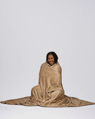 Woman sitting on the floor wrapped in a tan oversized blanket, smiling against a neutral studio background.