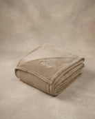 Folded tan oversized blanket with embroidered logo displayed on a matching neutral backdrop.