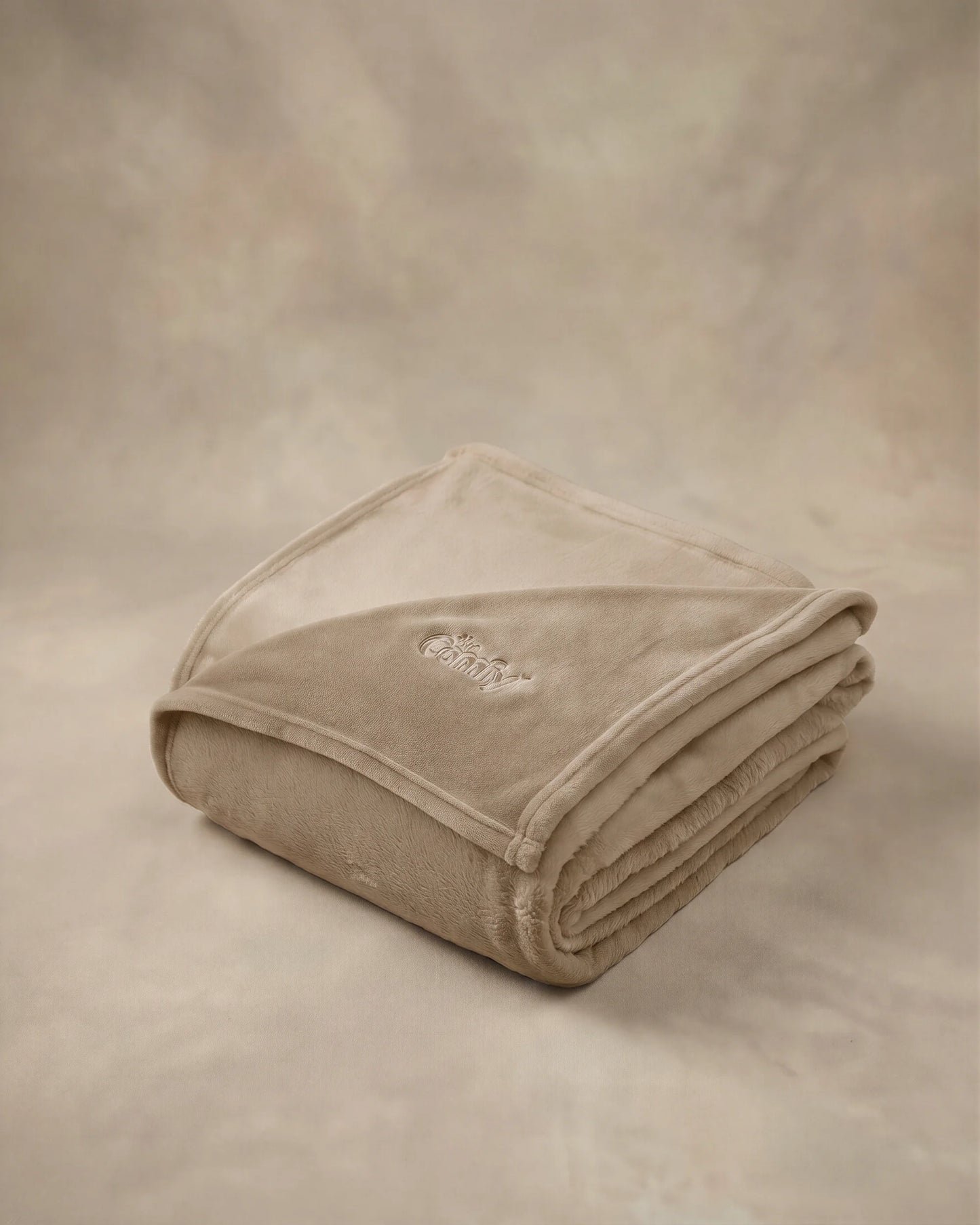 Folded tan oversized blanket with embroidered logo displayed on a matching neutral backdrop.