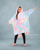 Girl standing with arms outstretched in a pastel tie-dye wearable blanket with a hood, against a light blue studio backdrop.
