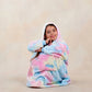 Girl sitting comfortably in a pastel tie-dye wearable blanket with her chin resting on her hand against a neutral studio background.