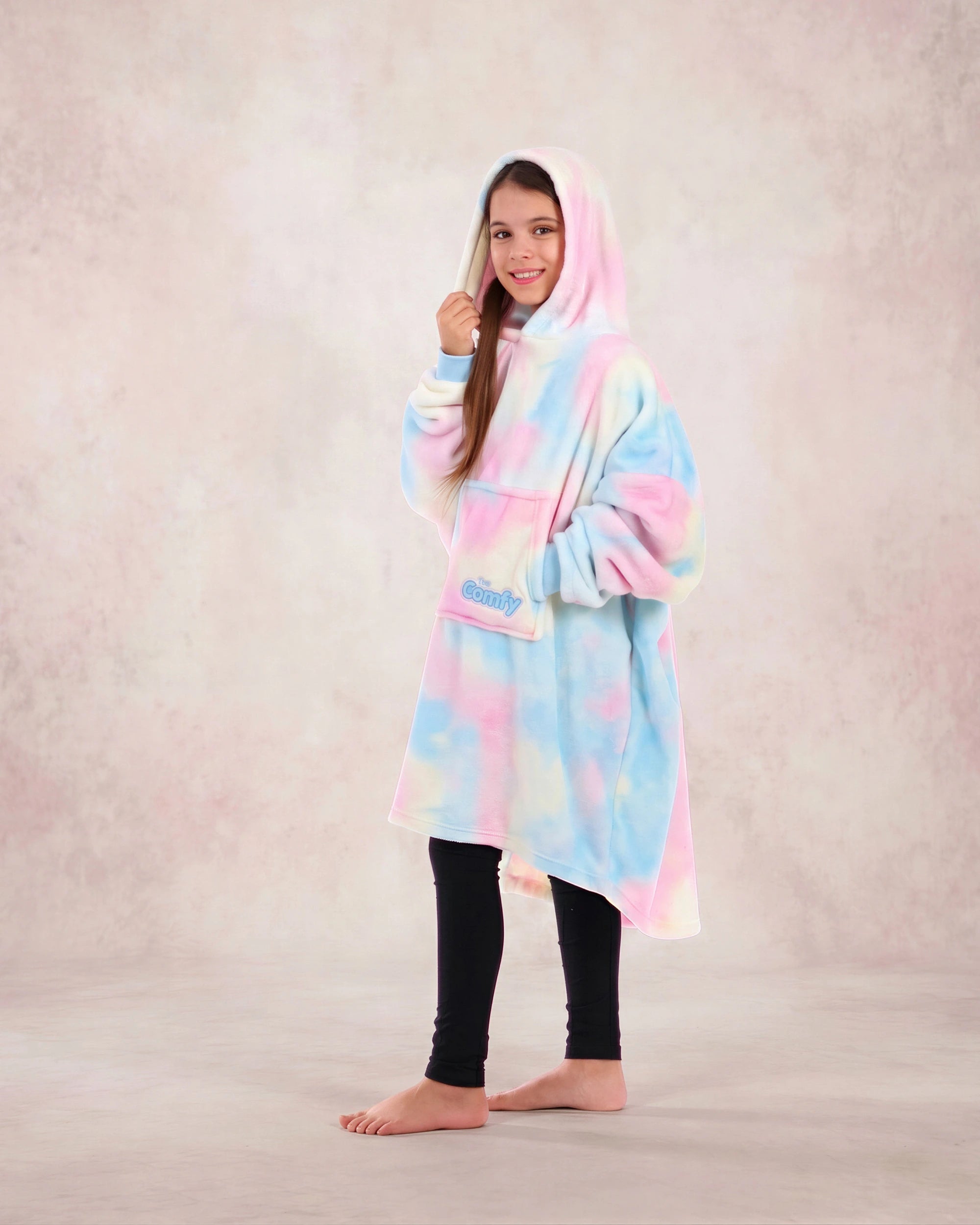 Girl standing in profile wearing a pastel tie-dye wearable blanket with a hood against a neutral studio backdrop.
