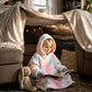 Child reading a book while wearing a pastel tie-dye wearable blanket inside a cozy blanket fort in a living room.