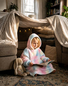 Child reading a book while wearing a pastel tie-dye wearable blanket inside a cozy blanket fort in a living room.