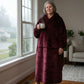 Woman standing by a window holding a mug while wearing The Dream Long in Burgundy in a cozy home setting.