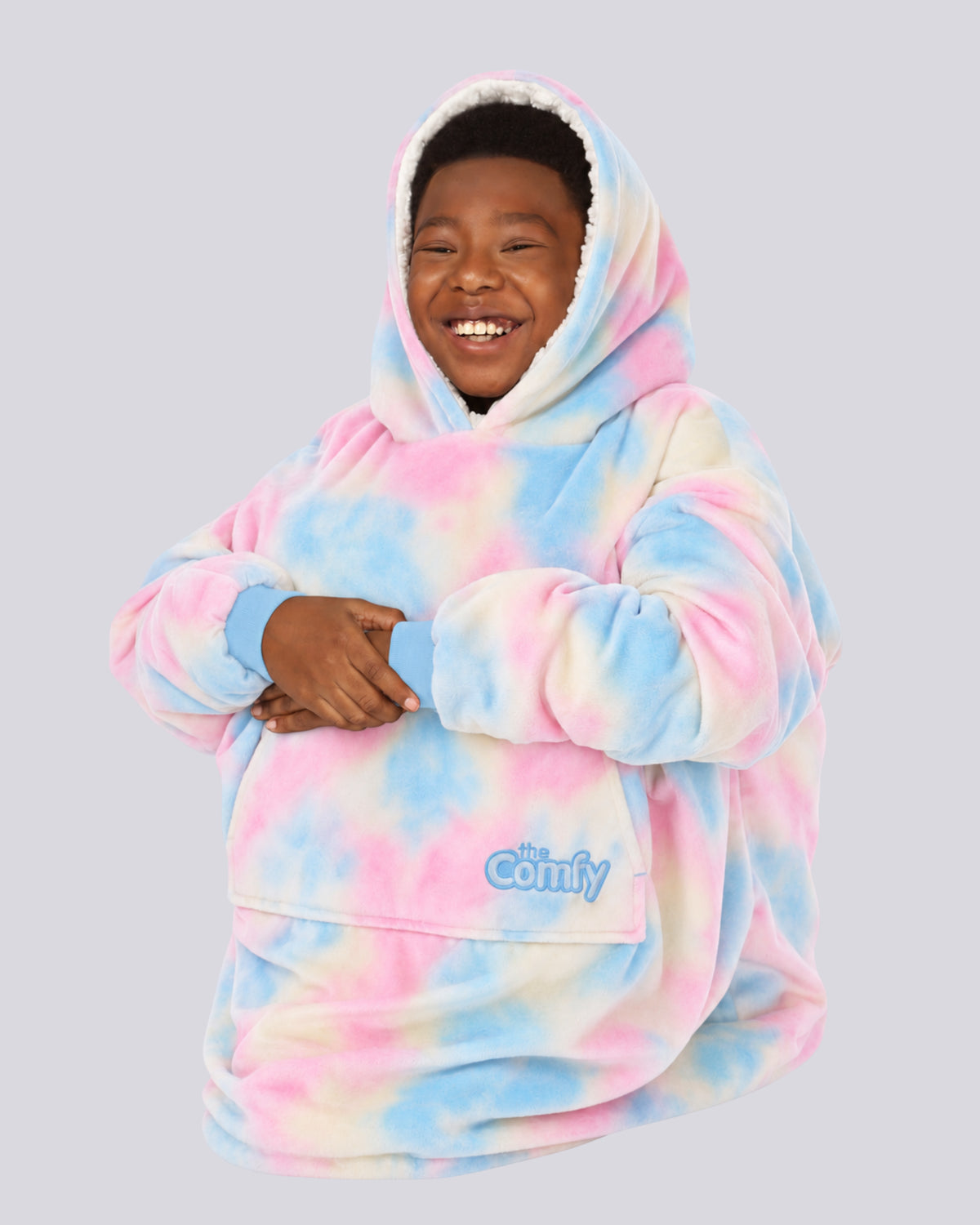 A smiling child sitting down curled up in a ball with the hood up of the Original Jr. wearable blanket in Cotton Candy Tie Dye.
