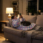 Child relaxing on a couch with a cat while wearing The Original Jr. Flower Checkered wearable blanket, illustrating everyday comfort for lounging and screen time.