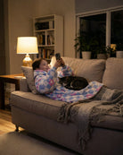 Child relaxing on a couch with a cat while wearing The Original Jr. Flower Checkered wearable blanket, illustrating everyday comfort for lounging and screen time.