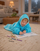 Child relaxing on the floor and coloring while wearing The Original Jr. Neon Aqua wearable blanket, perfect for cozy mornings, quiet activities, and comfort at home.