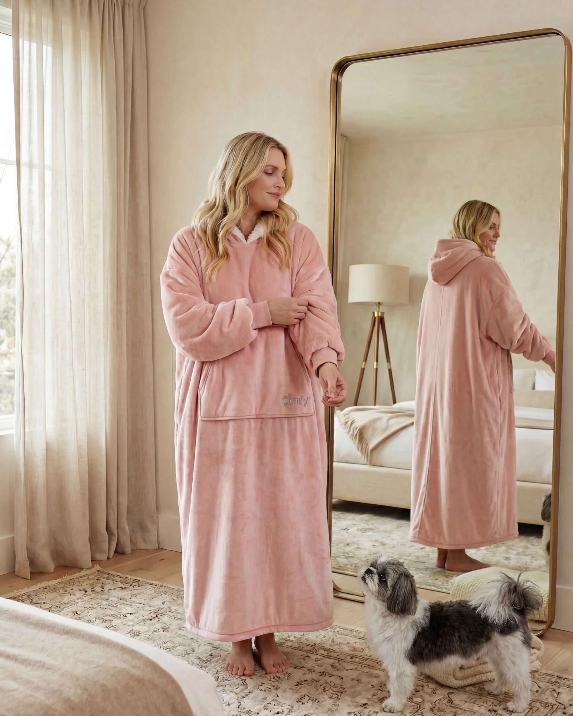 Woman wearing The Comfy Original Long in blush in a cozy bedroom, standing beside a mirror with her dog.