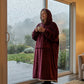 Woman wearing The Comfy Original Long in burgundy while enjoying coffee indoors beside a rainy window.