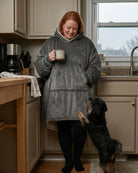 Woman wearing The Original wearable blanket in charcoal holding a mug in a kitchen with a dog nearby