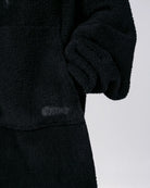 Close-up of The Teddy Bear Quarter-Zip wearable blanket in black showing sleeve detail and embroidered logo on CloudSherpa™ fabric