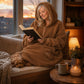 Lifestyle view of The Teddy Bear Quarter-Zip wearable blanket in camel worn while sitting by a window reading with a dog, showing cozy at-home use