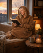 Lifestyle view of The Teddy Bear Quarter-Zip wearable blanket in camel worn while sitting by a window reading with a dog, showing cozy at-home use