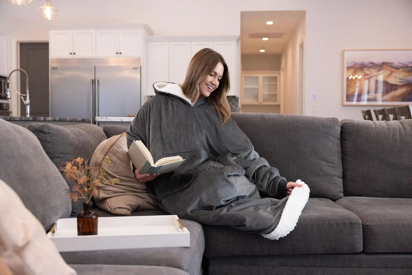 Person wearing an original long gray the comfy and reading a book on a couch in a modern living room.