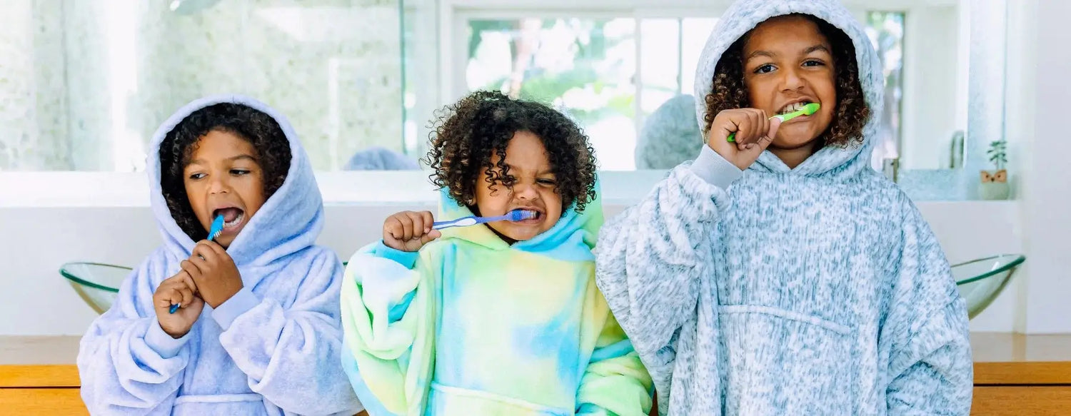 three kids with curly hair wearing  wearable blankets from The Comfy and brushing their teeth