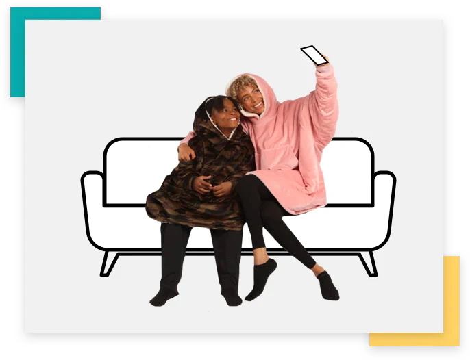 Two people sitting in a couch, taking a selfie together and wearing wearable blankets from The Comfy